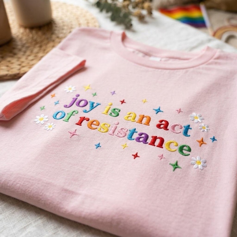 May include: A light pink t-shirt with the embroidered phrase "joy is an act of resistance" in rainbow colors. The design includes small embroidered flowers and stars.