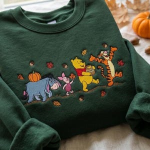 May include: A dark green crewneck sweatshirt featuring an embroidered design of Eeyore with a pumpkin, Piglet, Winnie the Pooh holding a honey pot, and Tigger. The scene includes scattered autumn leaves and acorns across the front of the garment.