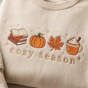 May include: A beige crewneck sweatshirt featuring embroidered designs of a stack of books, an orange pumpkin, a maple leaf, and a mug with whipped cream and a cinnamon stick. The text "cozy season" is embroidered below with small star accents.