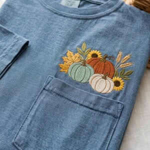May include: A blue t-shirt with a chest pocket featuring an embroidered design of three pumpkins in light green, orange, and white, surrounded by yellow sunflowers, golden wheat stalks, and autumn leaves in shades of yellow and green.
