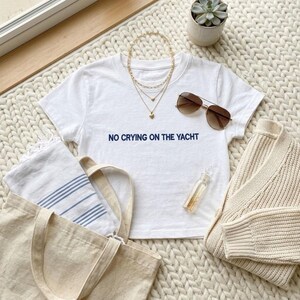 Puede incluir: Una composici&oacute;n plana con una camiseta blanca con el texto en azul marino "NO CRYING ON THE YACHT", collares dorados superpuestos, gafas de sol, un su&eacute;ter color crema, un bolso de mano y una peque&ntilde;a suculenta en una maceta blanca.