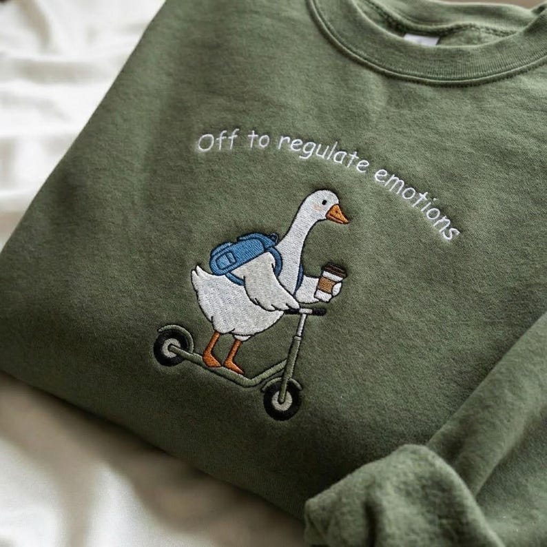 Puede incluir: Sudadera verde oliva con un dise&ntilde;o bordado de un ganso en patinete, con mochila y taza de caf&eacute;. El texto "Off to regulate emotions" est&aacute; arqueado sobre el ganso. La sudadera es de material suave.