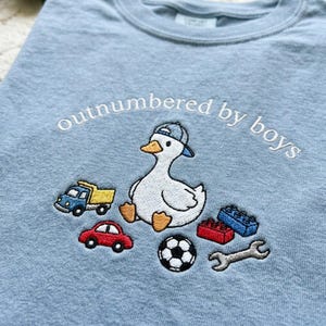 K&ouml;nnte beinhalten: Hellblaues T-Shirt mit dem gestickten Schriftzug "outnumbered by boys". Das Design zeigt eine gestickte Ente mit Kappe, umgeben von Spielzeugautos, Bausteinen, einem Fu&szlig;ball und einem Schraubenschl&uuml;ssel.