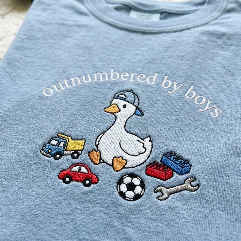 K&ouml;nnte beinhalten: Hellblaues T-Shirt mit dem gestickten Schriftzug "outnumbered by boys". Das Design zeigt eine gestickte Ente mit Kappe, umgeben von Spielzeugautos, Bausteinen, einem Fu&szlig;ball und einem Schraubenschl&uuml;ssel.