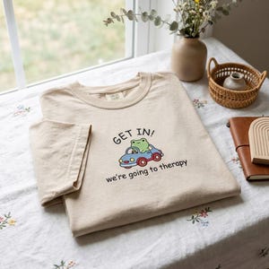 May include: Beige t-shirt with a crew neck. The shirt features a cartoon frog driving a blue car with the text "GET IN! we're going to therapy". The shirt is folded on a floral tablecloth.