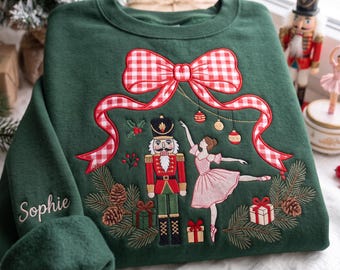 May include: A dark green crewneck sweatshirt featuring an embroidered design of a nutcracker and a ballerina surrounded by pine branches, gifts, and a large red and white gingham bow. The name Sophie is embroidered on the left sleeve.