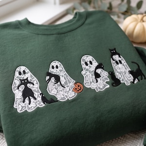 Könnte beinhalten: Ein dunkelwaldgrüner Rundhals-Pullover mit einer Stickerei von vier weißen Geistern im Spitzenmuster. Die Geister werden von mehreren schwarzen Katzen in verschiedenen Posen begleitet, und ein kleiner orangefarbener Kürbis ist abgebildet.