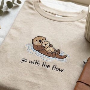 Op de afbeelding: Beige T-shirt met een geborduurd ontwerp van twee otters die in het water drijven. De grotere otter omarmt een kleinere. De tekst "go with the flow" is eronder in het zwart geborduurd.