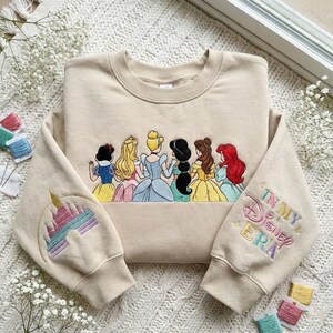 May include: Beige sweatshirt featuring an embroidered design of Disney princesses and a castle on the sleeve. The right sleeve has the text "In My Disney Era" in pastel colors. The princesses are depicted in their iconic gowns.
