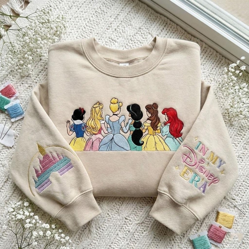 May include: Beige sweatshirt featuring an embroidered design of Disney princesses and a castle on the sleeve. The right sleeve has the text "In My Disney Era" in pastel colors. The princesses are depicted in their iconic gowns.