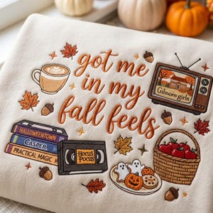 May include: A cream-colored sweatshirt featuring embroidered autumn-themed designs and the text "got me in my fall feels." The embroidery includes a coffee mug, a vintage television displaying "Gilmore girls," a stack of VHS tapes, a basket of apples, and cookies.
