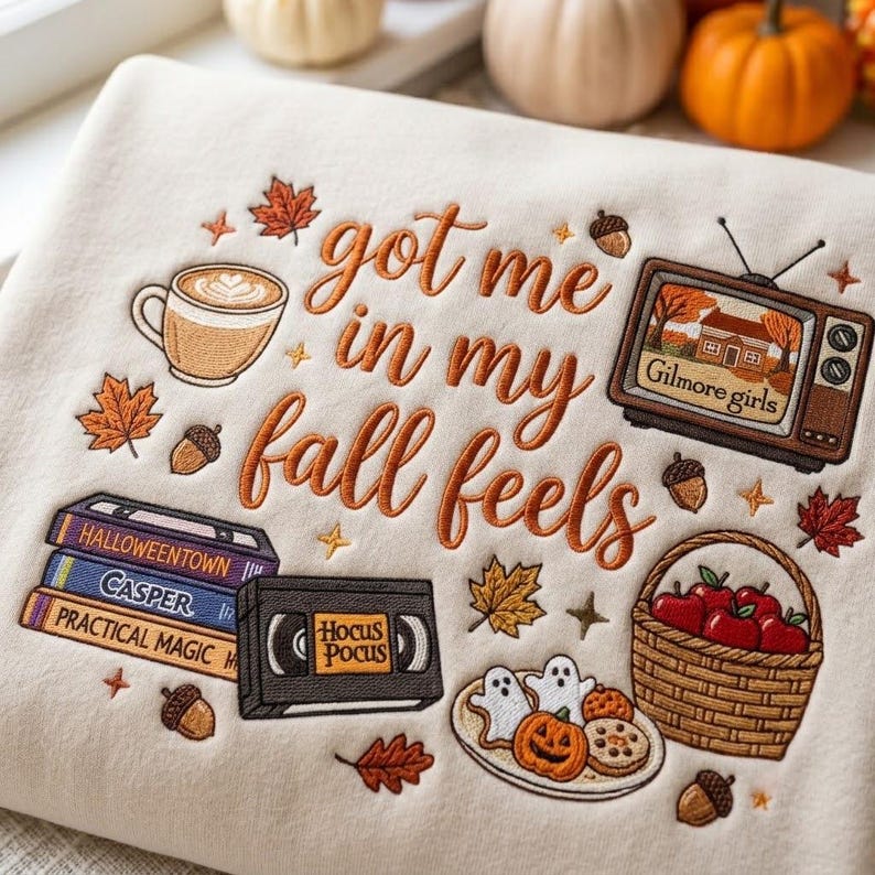 May include: A cream-colored sweatshirt featuring embroidered autumn-themed designs and the text "got me in my fall feels." The embroidery includes a coffee mug, a vintage television displaying "Gilmore girls," a stack of VHS tapes, a basket of apples, and cookies.