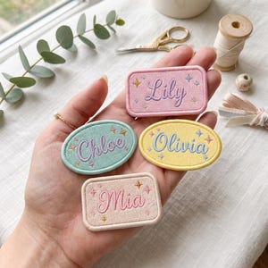 May include: Four embroidered fabric patches in rectangular and oval shapes. The patches feature the names Lily, Chloe, Olivia, and Mia in cursive script, accented with small embroidered star motifs in various pastel colors including pink, blue, and yellow.