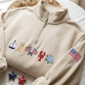 May include: Cream-colored sweatshirt with a quarter-zip neckline and embroidered nautical and patriotic designs. Features a sailboat, golden retriever, American flags, lobster, and a blue flower. An American flag patch is on the sleeve.