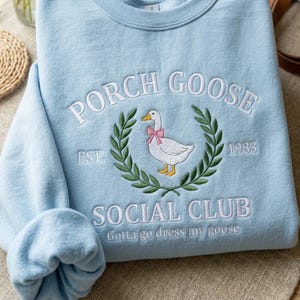 May include: Pale blue sweatshirt featuring embroidered text and a goose design. The text says "PORCH GOOSE SOCIAL CLUB" with a goose wearing a pink bow, encircled by a green wreath. The sweatshirt also has the text "EST. 1983" and "Gotta go dress my goose".