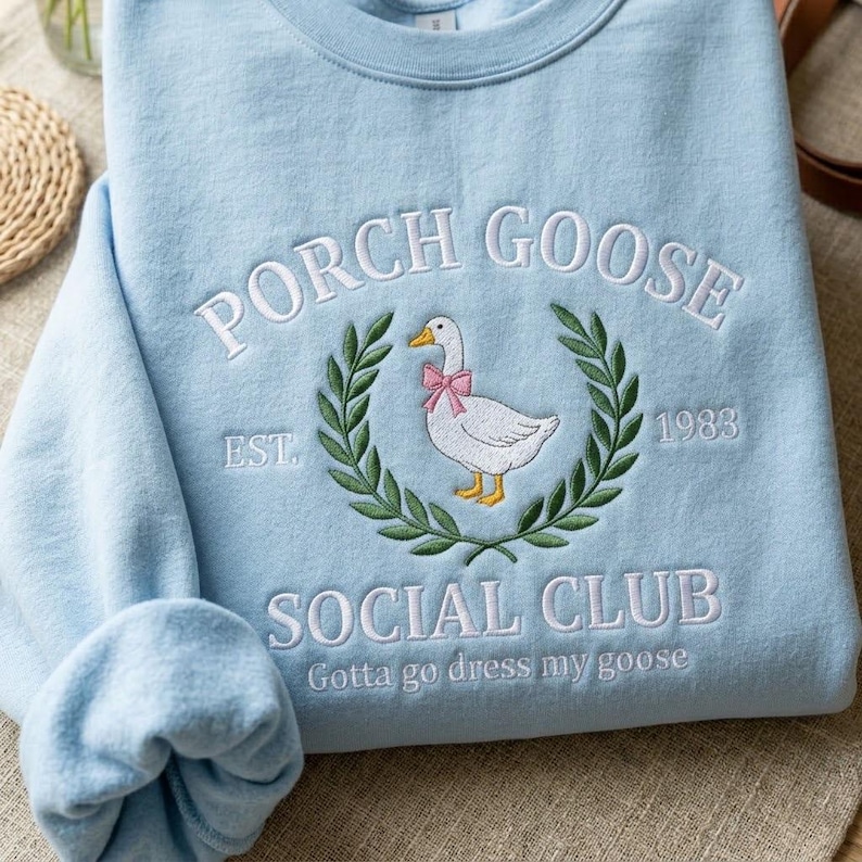 May include: Pale blue sweatshirt featuring embroidered text and a goose design. The text says "PORCH GOOSE SOCIAL CLUB" with a goose wearing a pink bow, encircled by a green wreath. The sweatshirt also has the text "EST. 1983" and "Gotta go dress my goose".