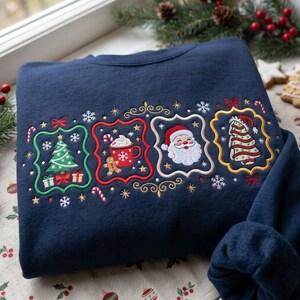 K&ouml;nnte beinhalten: Ein marineblaues Sweatshirt mit gestickten Weihnachtsmotiven in vier dekorativen Rahmen. Die Designs zeigen einen gr&uuml;nen Weihnachtsbaum, eine rote Tasse mit hei&szlig;er Schokolade und Lebkuchenmann, ein Weihnachtsmann-Gesicht und einen Baum.