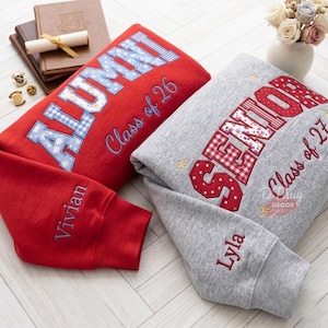 Puede incluir: Una sudadera roja con letras de aplique a cuadros azules que forman ALUMNI y el texto Class of 26, y una sudadera gris con letras de aplique a cuadros rojos que forman SENIOR y el texto Class of 27, ambas con nombres bordados en las mangas.