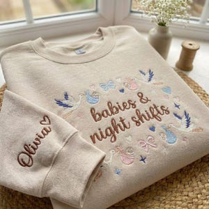 May include: Beige sweatshirt with embroidered text "babies & night shifts" and the name "Olivia" on the sleeve. The design includes storks, babies, bows, and stars in blue, pink, and beige.