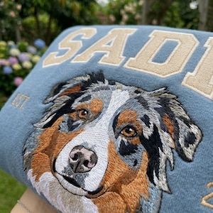 May include: A blue embroidered pillow featuring a detailed portrait of a dog with brown, black, and white fur. The name "SADIE" is embroidered in tan block letters across the top, with "EST." below. The dog's eyes are brown.