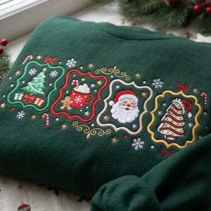 K&ouml;nnte beinhalten: Ein dunkelgr&uuml;nes Sweatshirt mit gestickten Weihnachtsmotiven in vier dekorativen Rahmen. Die Designs zeigen einen Weihnachtsbaum mit Geschenken, eine Tasse Kakao mit Lebkuchenmann, ein Gesicht vom Weihnachtsmann und einen gestreiften Strumpf.