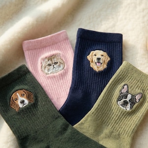 Könnte beinhalten: Vier Paar Socken in verschiedenen Farben, jede mit einem anderen Tiergesicht bestickt. Die Socken sind in Grün-, Rosa- und Marineblautönen gehalten und zeigen eine Katze, einen Beagle, einen Golden Retriever und eine französische Bulldogge.