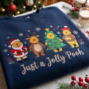Può includere: Una felpa blu navy con personaggi di Winnie the Pooh ricamati in abiti festivi, tra cui un costume da Babbo Natale, corna di renna, un costume da albero di Natale e un maglione invernale. Il testo "Just a Jolly Pooh" è ricamato sotto.