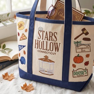 May include: A cream-colored canvas tote bag with navy blue trim and handles. The front features embroidered designs including a gazebo, a pumpkin, a gavel, and signs for Luke's, Dragonfly Inn, and Doose's Market, with text reading Stars Hollow Connecticut.