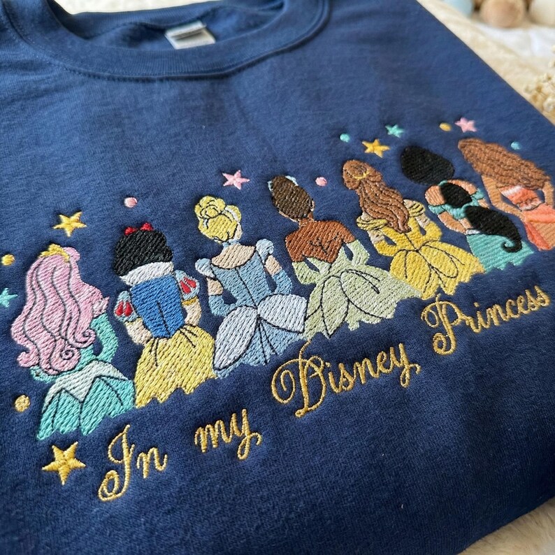 May include: Navy blue sweatshirt with embroidered Disney Princesses and the text "In my Disney Princess". The design features several princesses with colorful dresses and hair, along with embroidered stars.