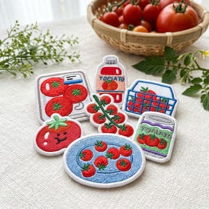 Op de afbeelding: Een verzameling geborduurde patches met tomatenthema's, waaronder een fles met het label TOMATO, een krat met tomaten, een blik met het label TOMATO, een tros tomaten, een bord met tomaten, een vierkante container en een glimlachende tomaat.