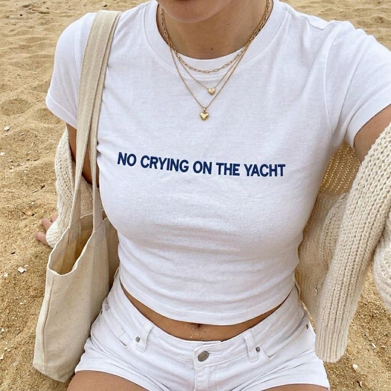 Puede incluir: Top corto blanco con el texto azul marino "NO CRYING ON THE YACHT". Se combina con shorts blancos y collares dorados superpuestos. Un bolso tote beige y un c&aacute;rdigan color crema completan el look.
