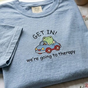 May include: Light blue t-shirt with the words "GET IN!" above an embroidered image of a green frog driving a blue car. The text "we're going to therapy" is below the car. The shirt has a crew neck and short sleeves.