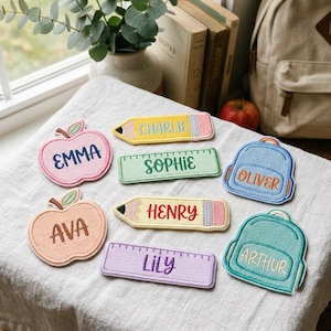 Pode incluir: Uma coleção de emblemas de tecido bordado com várias formas, incluindo maçãs, lápis, réguas e mochilas. Cada emblema apresenta um nome único costurado com fio colorido: Emma, Charlie, Sophie, Oliver, Ava, Henry e Lily.