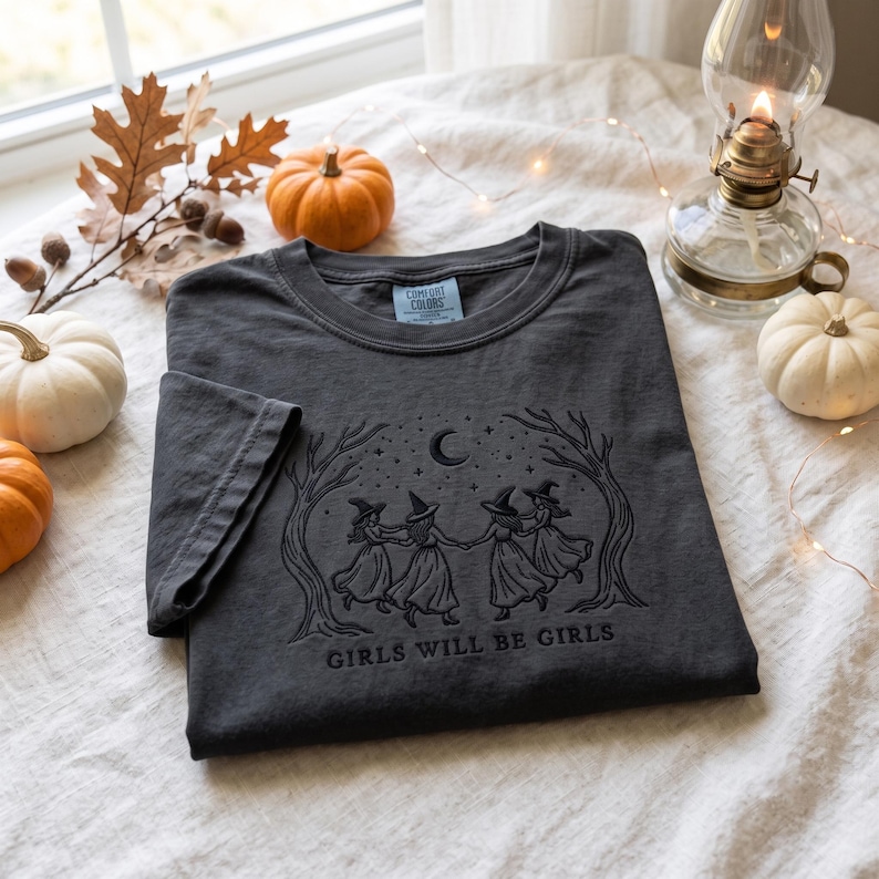 Peut inclure: Un t-shirt gris anthracite &agrave; manches courtes avec une broderie noire repr&eacute;sentant quatre silhouettes portant des chapeaux de sorci&egrave;re dansant en cercle sous un croissant de lune et des &eacute;toiles, encadr&eacute;es par deux arbres, avec le texte "GIRLS WILL BE GIRLS".