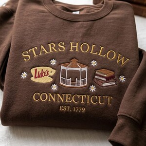 May include: A dark brown crewneck sweatshirt featuring gold embroidered text that reads STARS HOLLOW CONNECTICUT EST. 1779. The design includes a central gazebo, a sign reading Luke's, a stack of three books, and small white daisy flowers.