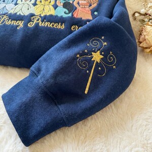 May include: Navy blue sweatshirt with embroidered Disney Princesses and the words "Disney Princess" in gold. The sleeve features a gold embroidered magic wand design. The sweatshirt is made of a soft material.