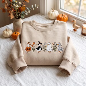 May include: A beige long-sleeve crewneck sweatshirt featuring an embroidered graphic of four ducks in costumes, including a pumpkin, a witch, a ghost, and a mummy, interspersed with small floral and botanical elements across the center chest.