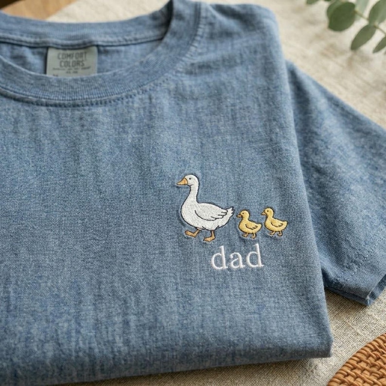 Puede incluir: Camiseta azul Comfort Colors con un dise&ntilde;o bordado de un pato blanco seguido de dos patitos amarillos. La palabra "dad" est&aacute; bordada debajo. La camiseta est&aacute; doblada sobre una superficie neutra.