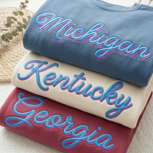 May include: A stack of three crewneck sweatshirts in blue, cream, and maroon. Each sweatshirt features embroidered cursive text in bright blue with a pink outline, displaying the names Michigan, Kentucky, and Georgia respectively.