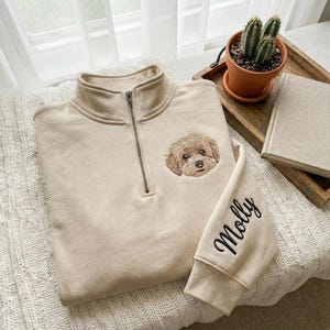 May include: A beige quarter-zip sweatshirt with a detailed embroidered dog face on the chest. The sleeve features the name "Molly" in black script. The sweatshirt is folded on a textured surface.
