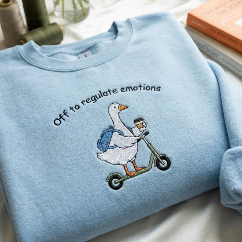 Puede incluir: Sudadera azul claro con un dise&ntilde;o bordado de un ganso montado en un patinete, sosteniendo una taza de caf&eacute; y llevando una mochila. El texto "Off to regulate emotions" est&aacute; bordado encima del ganso. La sudadera es de material suave.