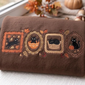 Puede incluir: Una sudadera marrón oscuro con cuatro marcos bordados rectangulares y ovalados. Cada marco contiene un gato negro en varias escenas otoñales, incluyendo un gato saltando entre hojas, sentado en un cuenco, durmiendo en una cesta y junto a una calabaza.