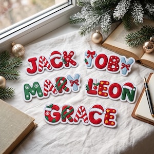 Può includere: Cinque targhette con nomi personalizzati a tema festivo. I nomi JACK, TOBY, MARY, LEON e GRACE sono visualizzati in lettere imbottite colorate, decorate con strisce di bastoncini di zucchero, cappelli di Babbo Natale, fiocchi, agrifoglio e stelle.