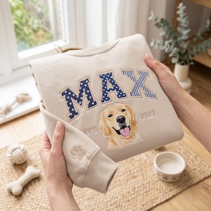 May include: A beige crewneck sweatshirt featuring the embroidered name MAX in blue and white patterned letters. Below the name is an embroidered golden retriever portrait with the text EST. 2023. A small embroidered paw print is on the sleeve cuff.