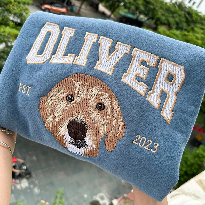 May include: A blue sweatshirt featuring the name "OLIVER" in white and gold embroidery. A detailed brown dog face is embroidered below the name, with the year "2023" beneath.