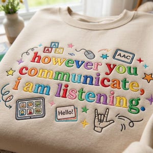 Op de afbeelding: Crèmekleurige sweatshirt met de geborduurde tekst "however you communicate, I am listening" in regenboogkleuren. Het ontwerp bevat een computermuis, sterren, een tablet en een handgebaar. Een comfortabel, casual kledingstuk.