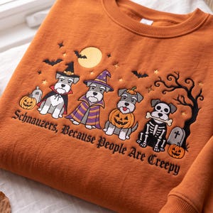 May include: An orange sweatshirt featuring an embroidered design of four Schnauzer dogs in Halloween costumes, including a witch, a wizard, a pumpkin, and a skeleton. The text reads, "Schnauzers, Because People Are Creepy," with bats, a moon, and tombstones.