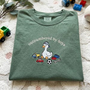 K&ouml;nnte beinhalten: Gr&uuml;nes T-Shirt mit dem gestickten Schriftzug "outnumbered by boys". Das Design zeigt eine Ente mit Baseballkappe, umgeben von Spielzeugautos, einem Fu&szlig;ball und Bausteinen. Das T-Shirt ist aus einem weichen, bequemen Material gefertigt.
