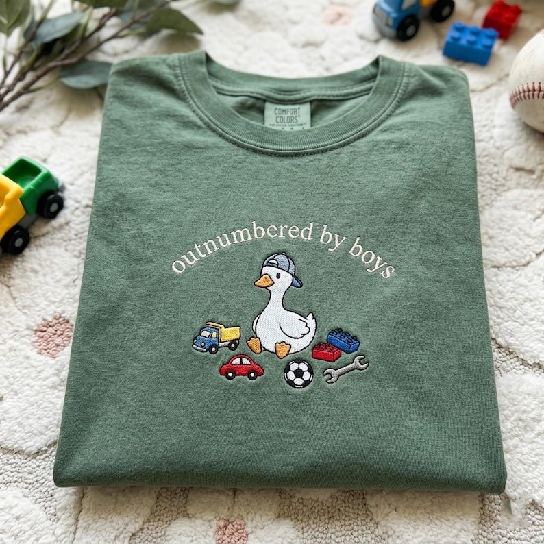 K&ouml;nnte beinhalten: Gr&uuml;nes T-Shirt mit dem gestickten Schriftzug "outnumbered by boys". Das Design zeigt eine Ente mit Baseballkappe, umgeben von Spielzeugautos, einem Fu&szlig;ball und Bausteinen. Das T-Shirt ist aus einem weichen, bequemen Material gefertigt.