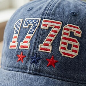 Puede incluir: Gorra de béisbol de mezclilla con el número "1776" bordado en rojo, blanco y azul, junto con tres estrellas bordadas. La gorra es de estilo clásico, perfecta para uso casual y eventos patrióticos.